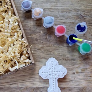 May include: A white ceramic cross with a Celtic knot design. The cross is surrounded by small paint pots in various colors, including brown, gold, gray, red, blue, and green.