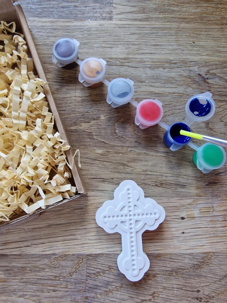 May include: A white ceramic cross with a Celtic knot design. The cross is surrounded by small paint pots in various colors, including brown, gold, gray, red, blue, and green.