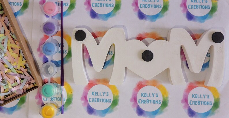 Pu&ograve; includere: Un cartello in legno bianco che scrive "MOM" con due magneti neri sul retro. Il cartello &egrave; circondato da barattoli di vernice colorati e un pennello.