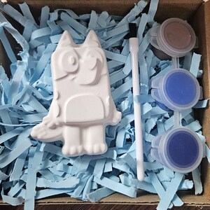 May include: A craft kit with a white cartoon dog figure, paint pots, and a paintbrush. The figure is surrounded by blue paper shreds in a cardboard box. The paint colors are blue and brown.