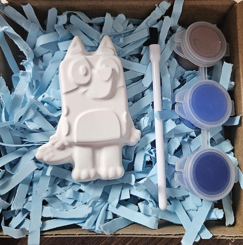 May include: A craft kit with a white cartoon dog figure, paint pots, and a paintbrush. The figure is surrounded by blue paper shreds in a cardboard box. The paint colors are blue and brown.