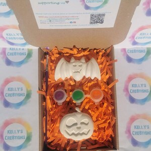 May include: A Halloween-themed craft kit in an open cardboard box. The kit includes a white bat and jack-o'-lantern, three paint pots, and a paintbrush, all nestled in orange paper shreds. The box has the text "KELLY'S CREATIONS".