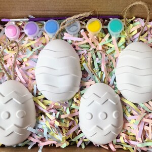 Puede incluir: Cuatro huevos de Pascua de cer&aacute;mica blanca con diferentes patrones, incluyendo l&iacute;neas onduladas y zigzags, est&aacute;n anidados en papel triturado de colores pastel. Los huevos est&aacute;n listos para ser pintados con las pinturas incluidas.