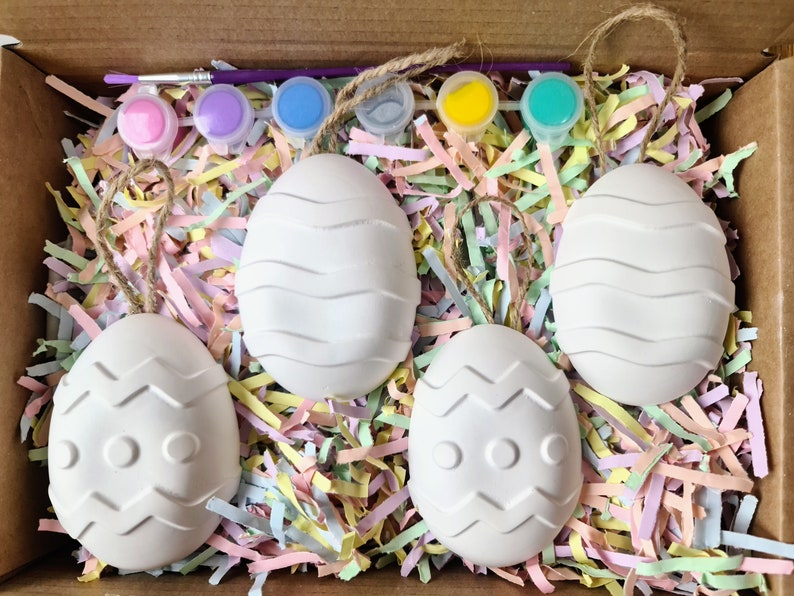 Puede incluir: Cuatro huevos de Pascua de cer&aacute;mica blanca con diferentes patrones, incluyendo l&iacute;neas onduladas y zigzags, est&aacute;n anidados en papel triturado de colores pastel. Los huevos est&aacute;n listos para ser pintados con las pinturas incluidas.