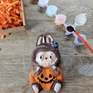 May include: A ceramic Halloween-themed figurine of a bunny wearing a pumpkin costume. The figurine is orange, brown, and white, with a smiling jack-o'-lantern face. Paint pots and a paintbrush are next to the figurine, suggesting a paint-your-own craft kit.