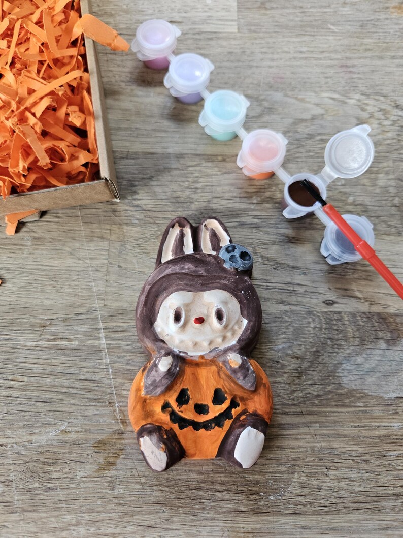 May include: A ceramic Halloween-themed figurine of a bunny wearing a pumpkin costume. The figurine is orange, brown, and white, with a smiling jack-o'-lantern face. Paint pots and a paintbrush are next to the figurine, suggesting a paint-your-own craft kit.