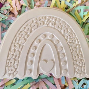 May include: Off-white, unfinished rainbow-shaped craft. The top layer features a leopard print design. The middle layer has a series of dots, and the bottom layer has a heart shape. A craft project for painting.