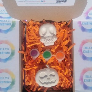May include: A Halloween-themed craft kit in an open cardboard box. The kit includes a white skull and jack-o'-lantern, three paint pots, and a paintbrush. The box is filled with orange paper shreds. The text "KELLY'S CREATIONS" is visible.
