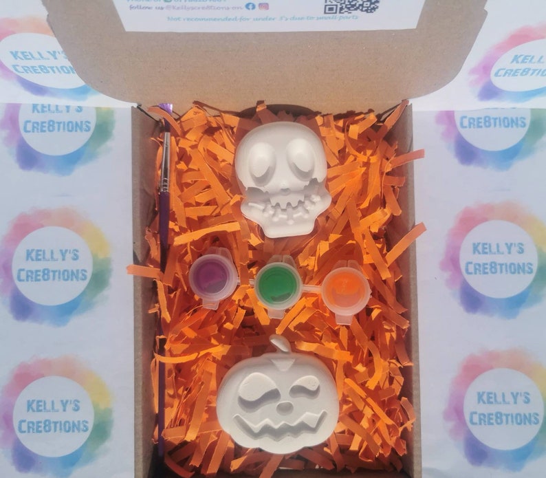 May include: A Halloween-themed craft kit in an open cardboard box. The kit includes a white skull and jack-o'-lantern, three paint pots, and a paintbrush. The box is filled with orange paper shreds. The text "KELLY'S CREATIONS" is visible.