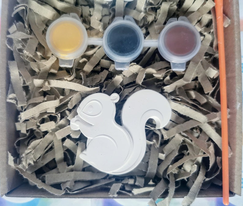 May include: A white ceramic squirrel figurine with a paint-by-numbers design. The kit includes three small paint pots with yellow, black, and brown paint.