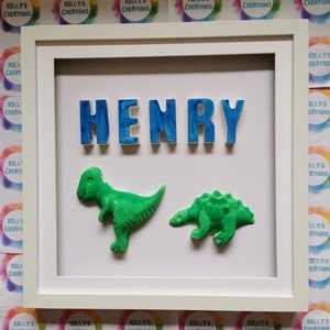 May include: A framed artwork with the name "HENRY" in blue, textured letters. Two green dinosaur figures are below the name. The white frame surrounds the artwork. The background is white. The artwork is a decorative piece.