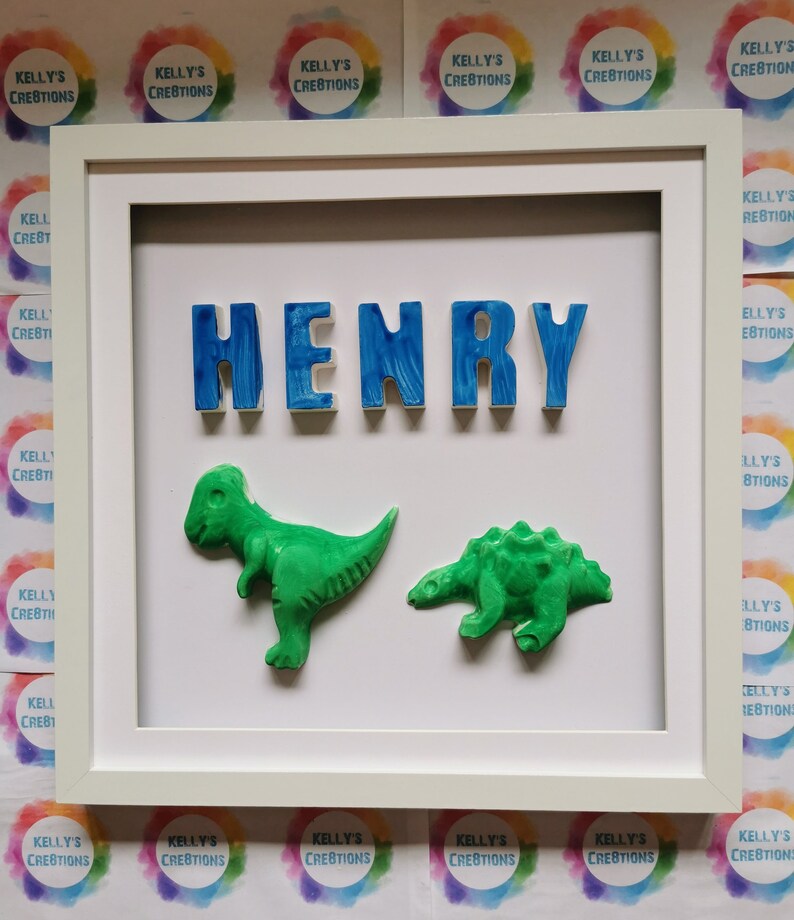May include: A framed artwork with the name "HENRY" in blue, textured letters. Two green dinosaur figures are below the name. The white frame surrounds the artwork. The background is white. The artwork is a decorative piece.