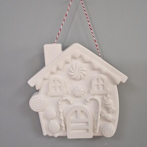 May include: White ceramic gingerbread house ornament with candy cane, swirl, and candy details. The ornament is hanging from a red and white twine.