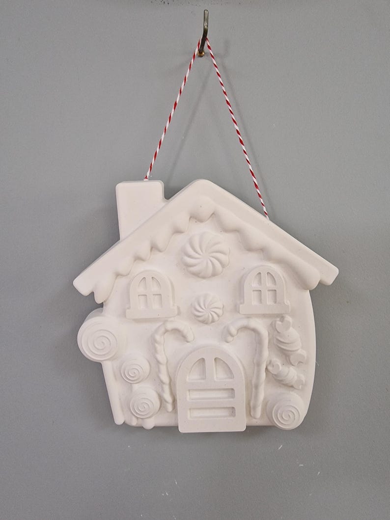 May include: White ceramic gingerbread house ornament with candy cane, swirl, and candy details. The ornament is hanging from a red and white twine.