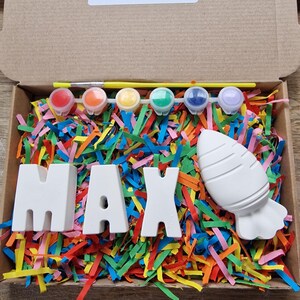 May include: A craft kit with the name MAX spelled out in white ceramic letters, a white ceramic rocket, and six small pots of paint. The kit is surrounded by colorful shredded paper.