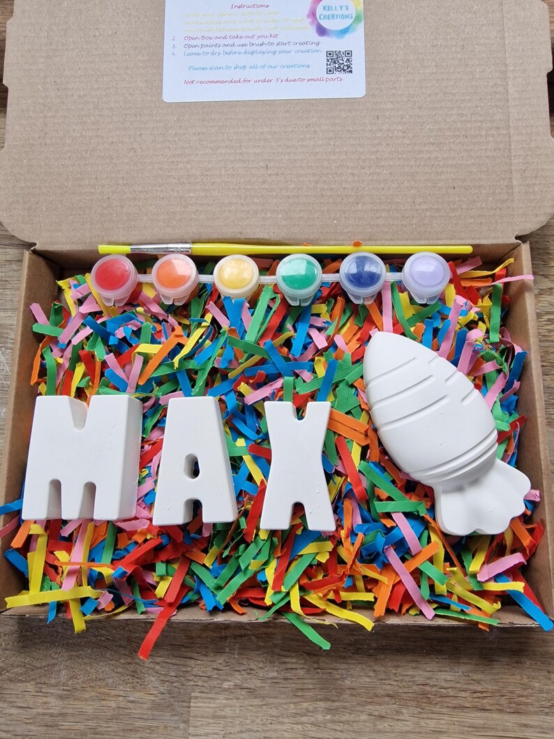 May include: A craft kit with the name MAX spelled out in white ceramic letters, a white ceramic rocket, and six small pots of paint. The kit is surrounded by colorful shredded paper.