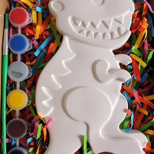 May include: A white ceramic dinosaur figurine in a box with colorful paper shreds and a set of paint pots and a paintbrush. The dinosaur has a toothy grin and is ready to be painted.