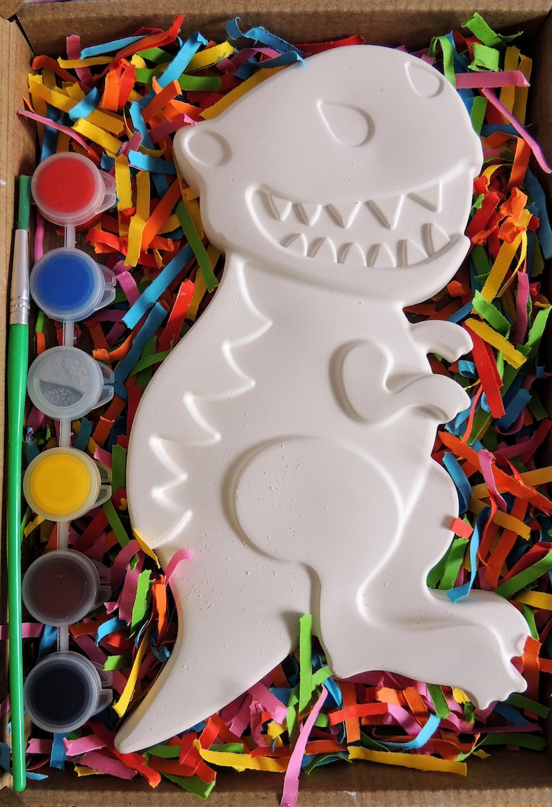 May include: A white ceramic dinosaur figurine in a box with colorful paper shreds and a set of paint pots and a paintbrush. The dinosaur has a toothy grin and is ready to be painted.