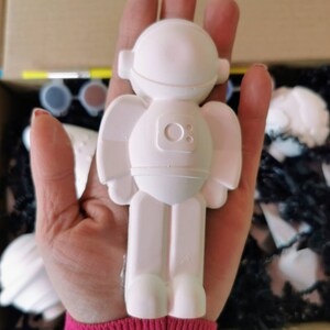 May include: White ceramic astronaut figurine with a round head and a small circle on the chest. The figurine is being held in a hand.