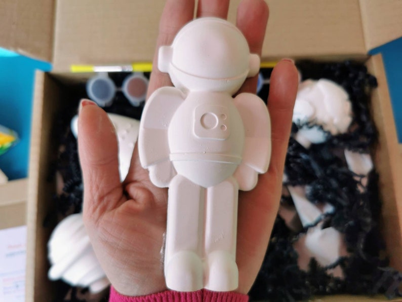 May include: White ceramic astronaut figurine with a round head and a small circle on the chest. The figurine is being held in a hand.