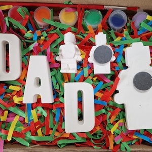 May include: A craft kit in a cardboard box, featuring the word "DAD" in white ceramic letters, along with paint pots, paintbrushes, and small ceramic figures. The box is filled with colorful shredded paper.