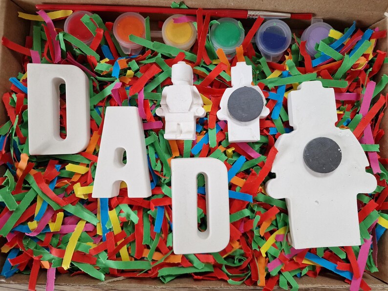 May include: A craft kit in a cardboard box, featuring the word "DAD" in white ceramic letters, along with paint pots, paintbrushes, and small ceramic figures. The box is filled with colorful shredded paper.
