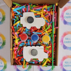 Puede incluir: Un kit de manualidades en una caja de cart&oacute;n, con dos imanes blancos con forma de cami&oacute;n, tres botes de pintura peque&ntilde;os (rojo, azul, amarillo) y un pincel verde. La caja est&aacute; llena de papel triturado colorido.