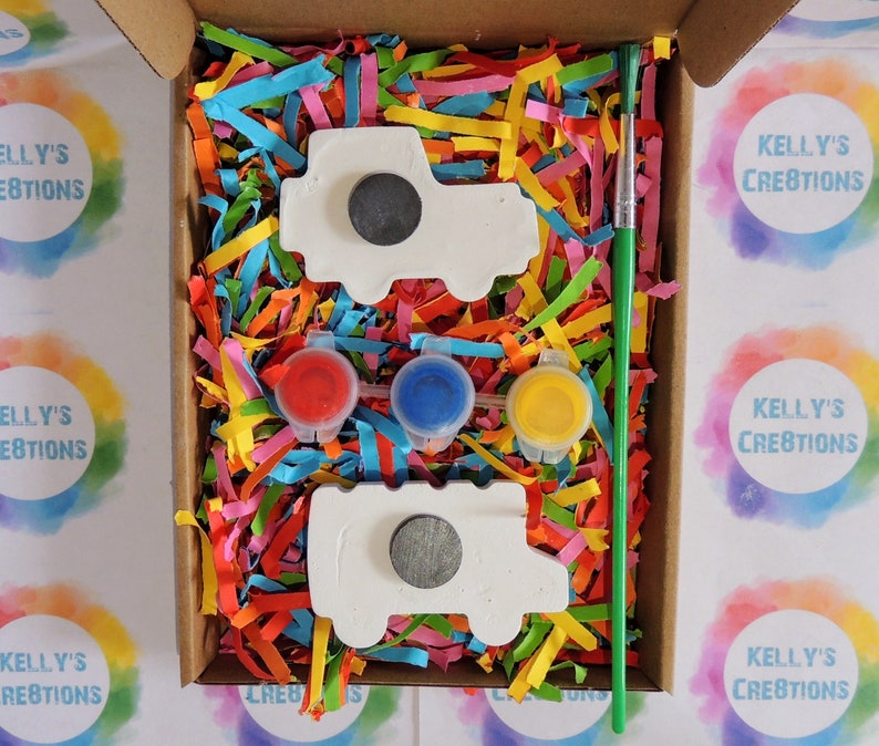 Puede incluir: Un kit de manualidades en una caja de cart&oacute;n, con dos imanes blancos con forma de cami&oacute;n, tres botes de pintura peque&ntilde;os (rojo, azul, amarillo) y un pincel verde. La caja est&aacute; llena de papel triturado colorido.