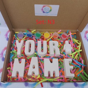 May include: A craft kit in a cardboard box, featuring the text "YOUR NAME" in large white letters, a small white figure, and six paint pots. The box is filled with colorful paper strips. The text "Set: N3" is visible.