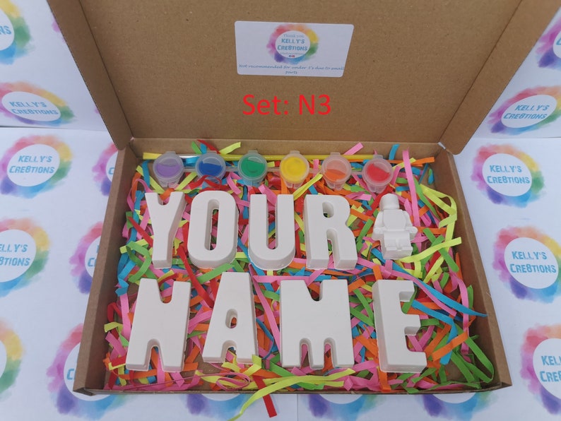 May include: A craft kit in a cardboard box, featuring the text "YOUR NAME" in large white letters, a small white figure, and six paint pots. The box is filled with colorful paper strips. The text "Set: N3" is visible.