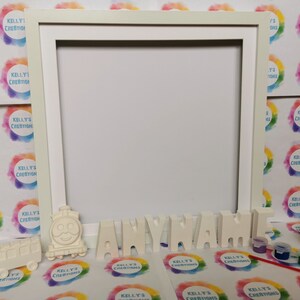 May include: A cream-coloured picture frame with the word "ANYNAME" in individual letters. A smiling train and bus are placed near the letters. Small paint pots and brushes are also visible, suggesting a DIY craft kit.
