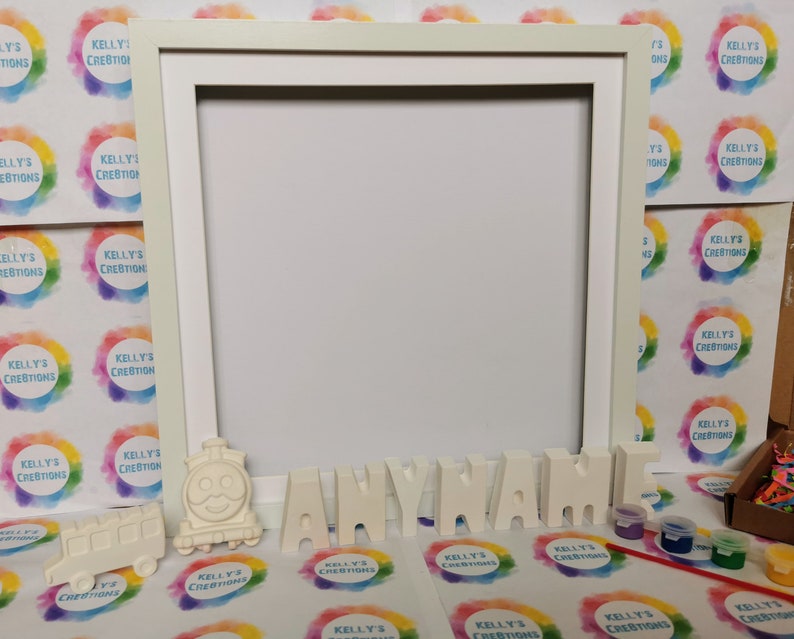 May include: A cream-coloured picture frame with the word "ANYNAME" in individual letters. A smiling train and bus are placed near the letters. Small paint pots and brushes are also visible, suggesting a DIY craft kit.