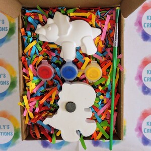 May include: A craft kit with two white ceramic dinosaur figurines, three small paint pots, a green paintbrush, and colorful shredded paper in a cardboard box. The box is labeled "Kelly's Creations".