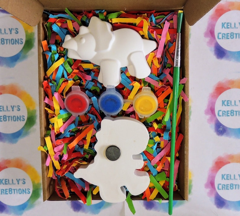 May include: A craft kit with two white ceramic dinosaur figurines, three small paint pots, a green paintbrush, and colorful shredded paper in a cardboard box. The box is labeled "Kelly's Creations".