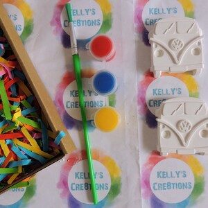 May include: A craft kit with two white ceramic Volkswagen bus figurines, a paintbrush, and four paint pots in red, blue, yellow, and white. The kit is packaged in a box with colorful shredded paper. The box has the text "Kelly's Creations" on it.
