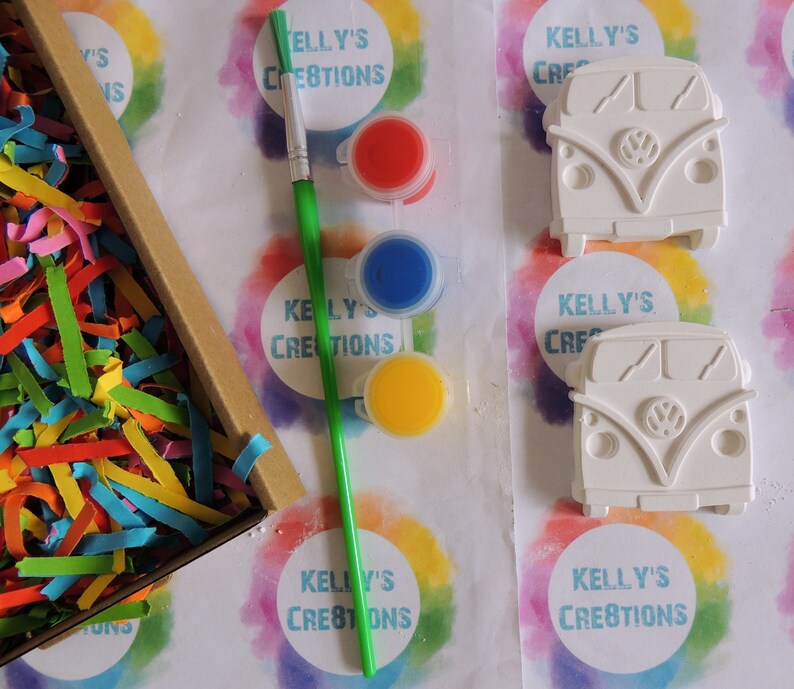 May include: A craft kit with two white ceramic Volkswagen bus figurines, a paintbrush, and four paint pots in red, blue, yellow, and white. The kit is packaged in a box with colorful shredded paper. The box has the text "Kelly's Creations" on it.