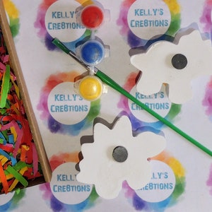 May include: Two white wooden flower-shaped ornaments with magnetic backs, paint pots in yellow, blue, and red, a paintbrush, and a green paint stirrer. The background is a white surface with colorful rainbow circles that say "Kelly's Creations".