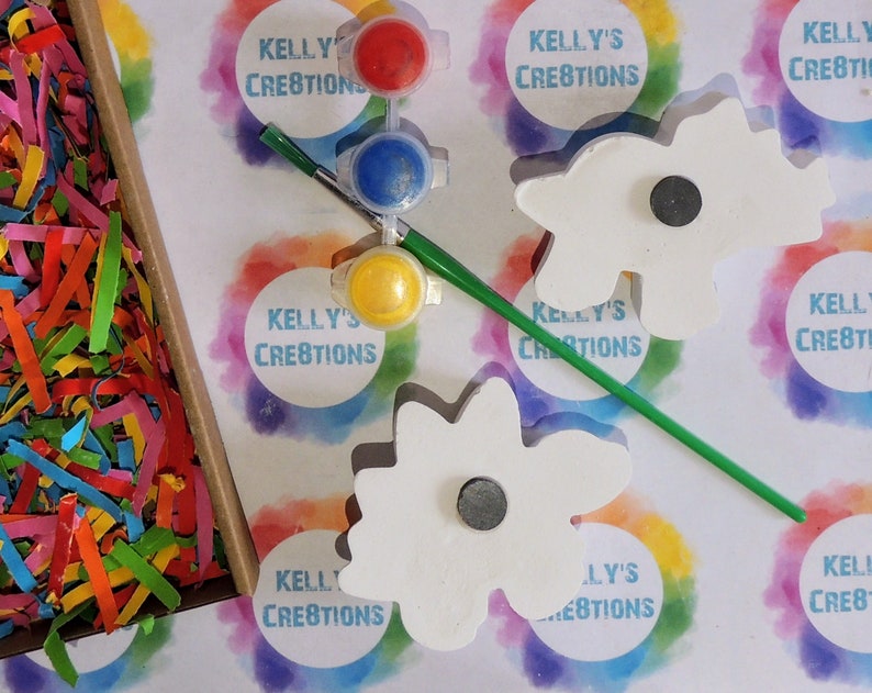 May include: Two white wooden flower-shaped ornaments with magnetic backs, paint pots in yellow, blue, and red, a paintbrush, and a green paint stirrer. The background is a white surface with colorful rainbow circles that say "Kelly's Creations".