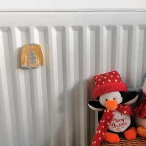 May include: Two penguin plush toys with red and white polka dot hats and scarves. The penguin on the left has the text "Merry Christmas" on its chest. The penguins are sitting on a wicker basket in front of a white radiator with a yellow ceramic snowman ornament.