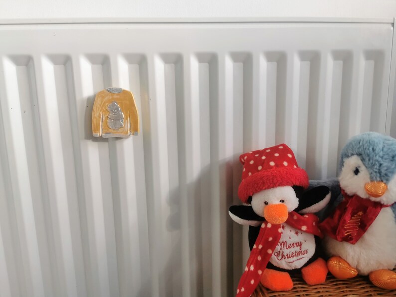 May include: Two penguin plush toys with red and white polka dot hats and scarves. The penguin on the left has the text "Merry Christmas" on its chest. The penguins are sitting on a wicker basket in front of a white radiator with a yellow ceramic snowman ornament.