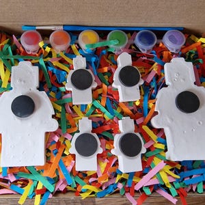 May include: A set of six white ceramic figurines with magnets on the back, ready to be painted. The figurines are in the shape of robots and are surrounded by colorful shredded paper. The set also includes five small paint pots with different colors and a paintbrush.