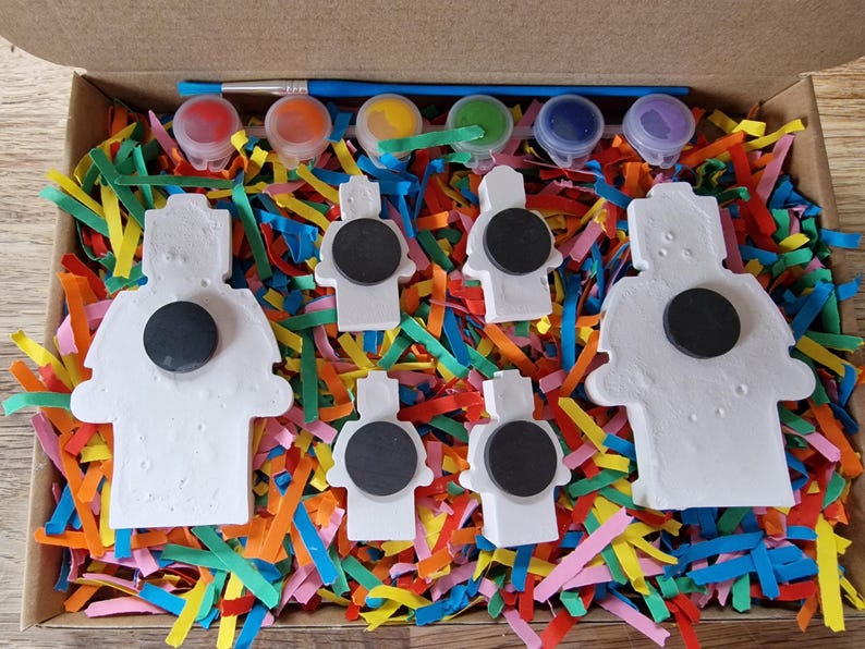 May include: A set of six white ceramic figurines with magnets on the back, ready to be painted. The figurines are in the shape of robots and are surrounded by colorful shredded paper. The set also includes five small paint pots with different colors and a paintbrush.