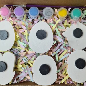 May include: A set of six white ceramic magnets in various shapes, including eggs, flowers, and stars. The magnets are ready to be painted with the included paints: pink, purple, blue, green, yellow, and a paintbrush. The magnets are packaged in a cardboard box with shredded paper.