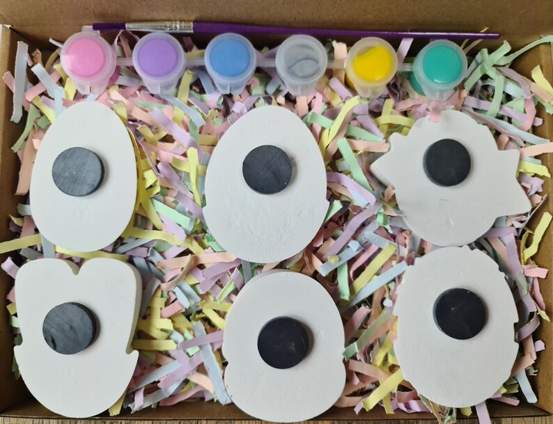 May include: A set of six white ceramic magnets in various shapes, including eggs, flowers, and stars. The magnets are ready to be painted with the included paints: pink, purple, blue, green, yellow, and a paintbrush. The magnets are packaged in a cardboard box with shredded paper.