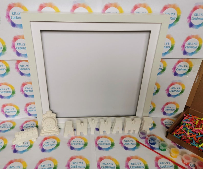 May include: A cream-colored picture frame with a train and the word "ANYNAME" in separate letters. Small paint pots, a paintbrush, and colorful paper strips are also included. The background features a repeating logo with the text "KELLY'S CREATIONS".