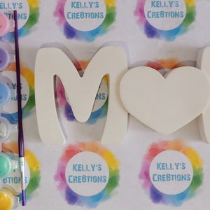 Pu&ograve; includere: Un kit fai-da-te per la creazione di un cartello in legno bianco "MAMMA" con un cuore, insieme a un set di barattoli di vernice e un pennello. I barattoli di vernice sono di vari colori, tra cui azzurro, verde, giallo e viola.