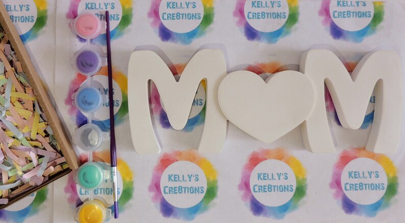 Pu&ograve; includere: Un kit fai-da-te per la creazione di un cartello in legno bianco "MAMMA" con un cuore, insieme a un set di barattoli di vernice e un pennello. I barattoli di vernice sono di vari colori, tra cui azzurro, verde, giallo e viola.