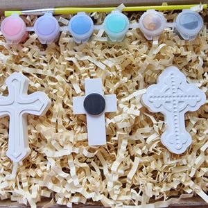May include: A set of three white ceramic crosses, two with a simple design and one with a more ornate design, in a box with paint pots and a brush. The paint pots are pink, light blue, blue, turquoise, and two clear pots.