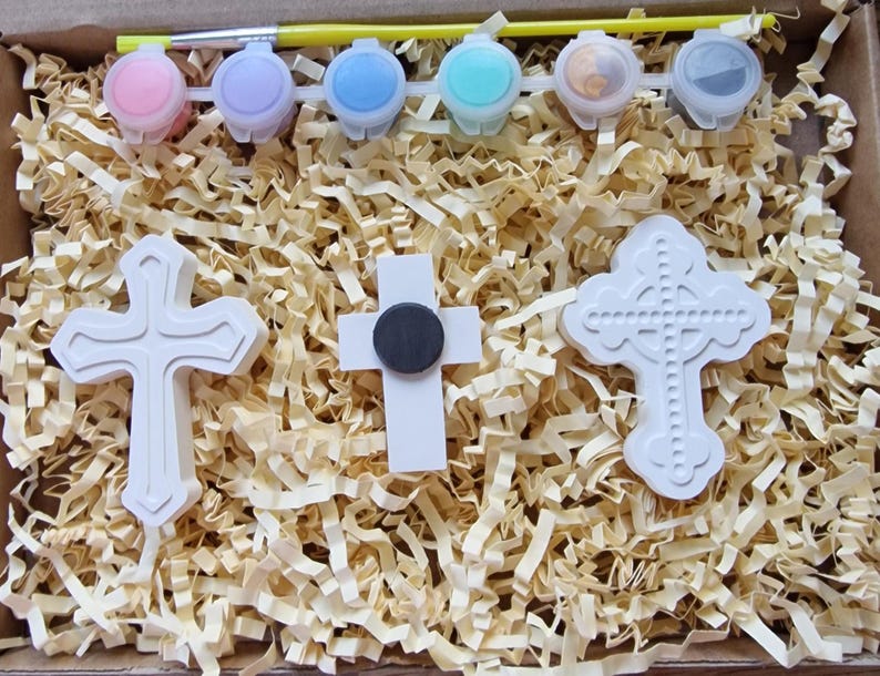 May include: A set of three white ceramic crosses, two with a simple design and one with a more ornate design, in a box with paint pots and a brush. The paint pots are pink, light blue, blue, turquoise, and two clear pots.