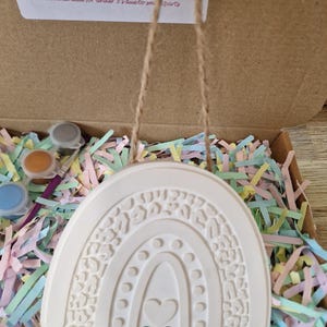 May include: A white ceramic rainbow ornament with a heart and leopard print design, hanging by a twine string. The ornament is in a cardboard box filled with colorful paper shreds, with paint pots and a brush visible.